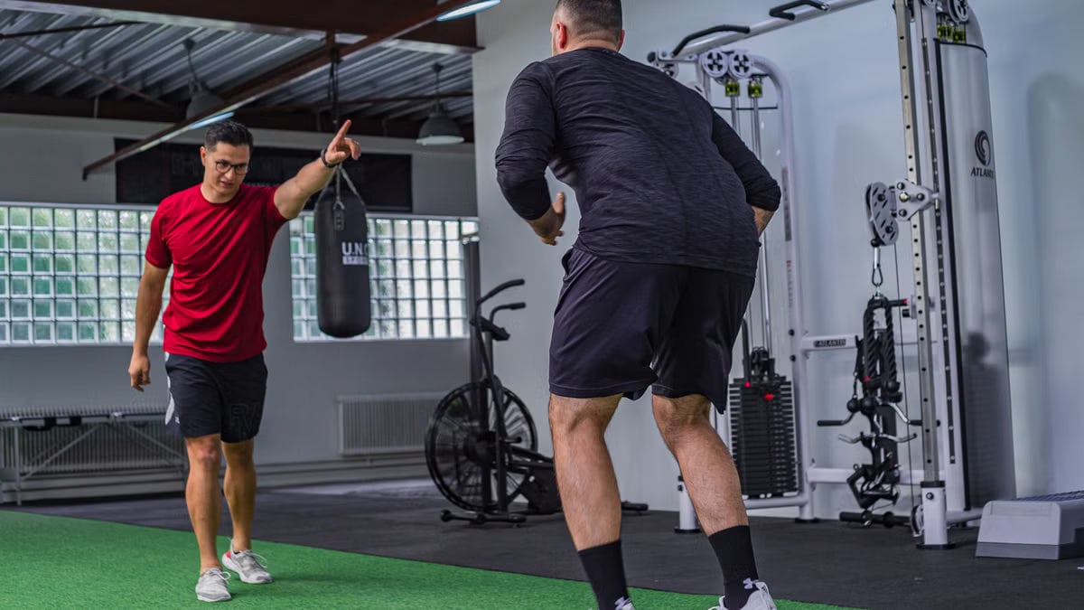 Back view of muscular man in sportswear doing exercises with trainer in modern fitness club
